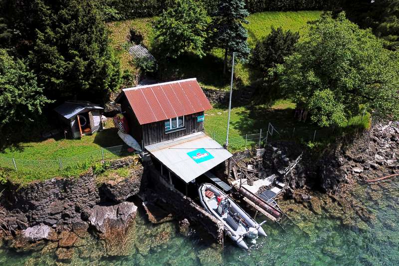 Die Luftaufnahme zeigt das charmante Bootshaus im Detail, inklusive privatem Anleger und dem bereitliegenden grauen Schlauchboot mit Außenbordmotor im glasklaren, schimmernden Wasser.