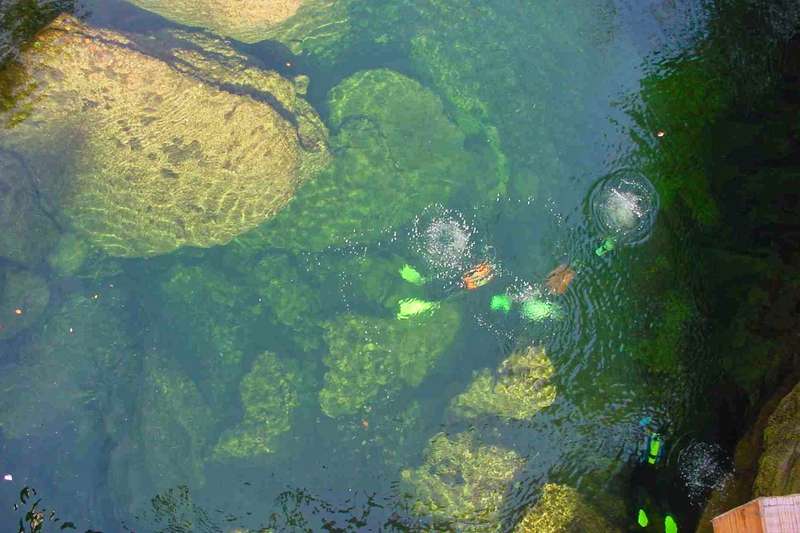 Blick von oben in glasklares Fluss-Wasser: Taucher erkunden eine faszinierende Unterwelt zwischen mächtigen Unterwasserfelsen. Die leuchtenden Flossen und aufsteigenden Luftblasen bilden einen farblichen Kontrast zum tiefen Grün des Gewässers.